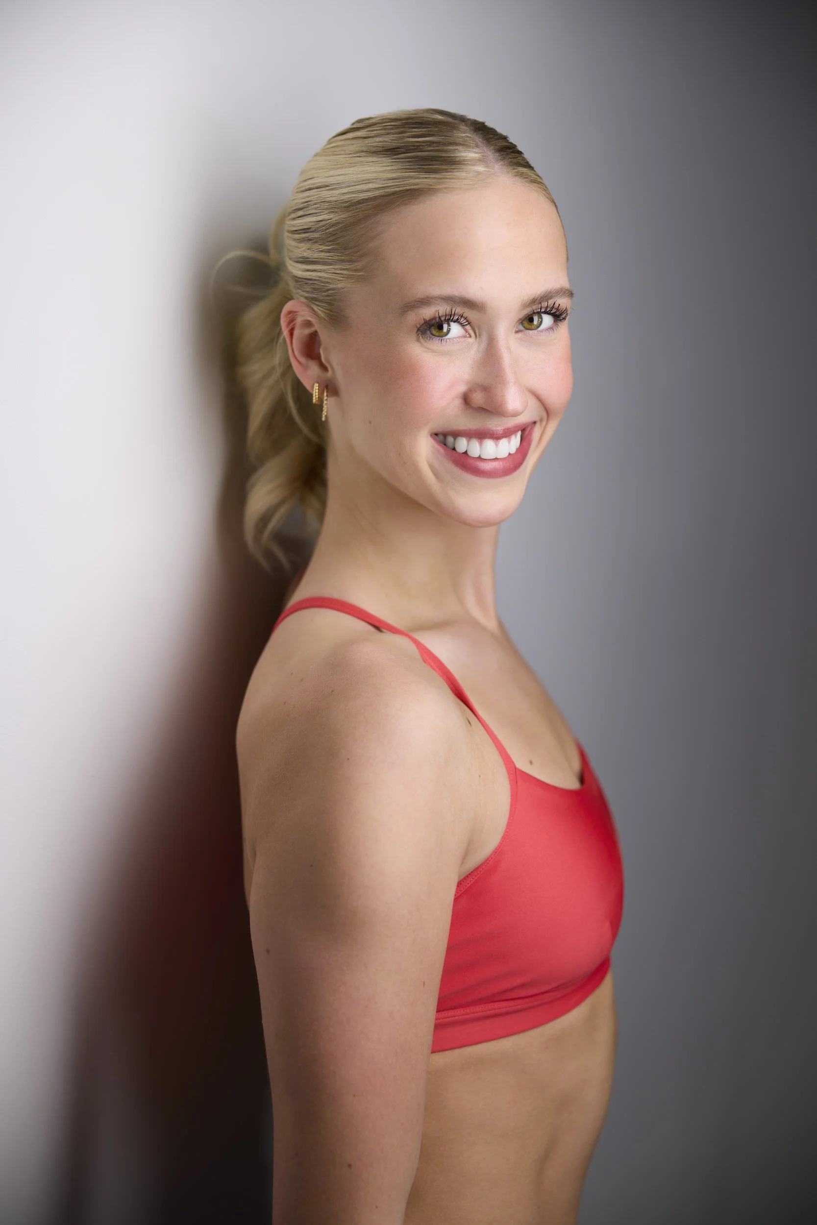 Woman with blonde hair in a ponytail, profile-turned smiling headshot in a red top against a grey background.