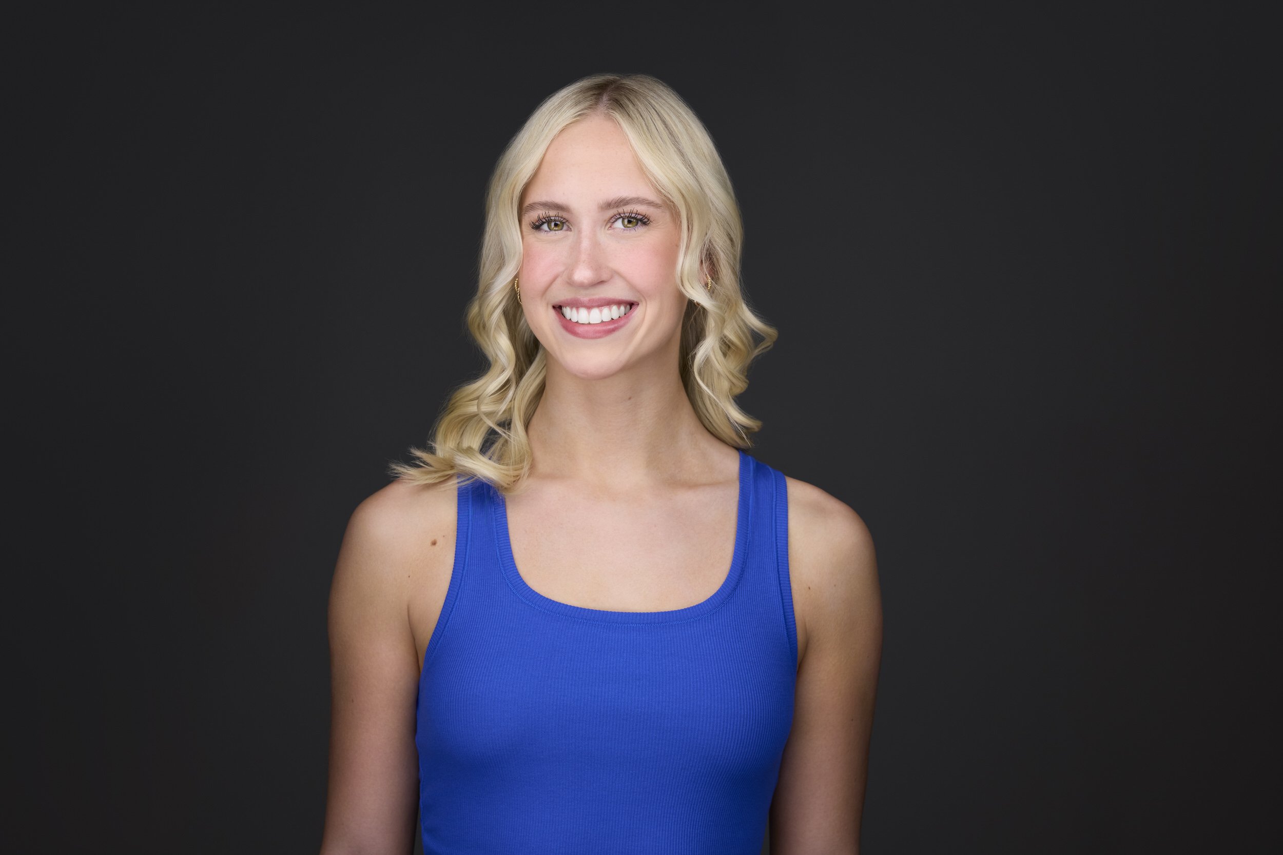 Woman with long blonde wavy hair in a blue tank top, smiling headshot against a dark background.