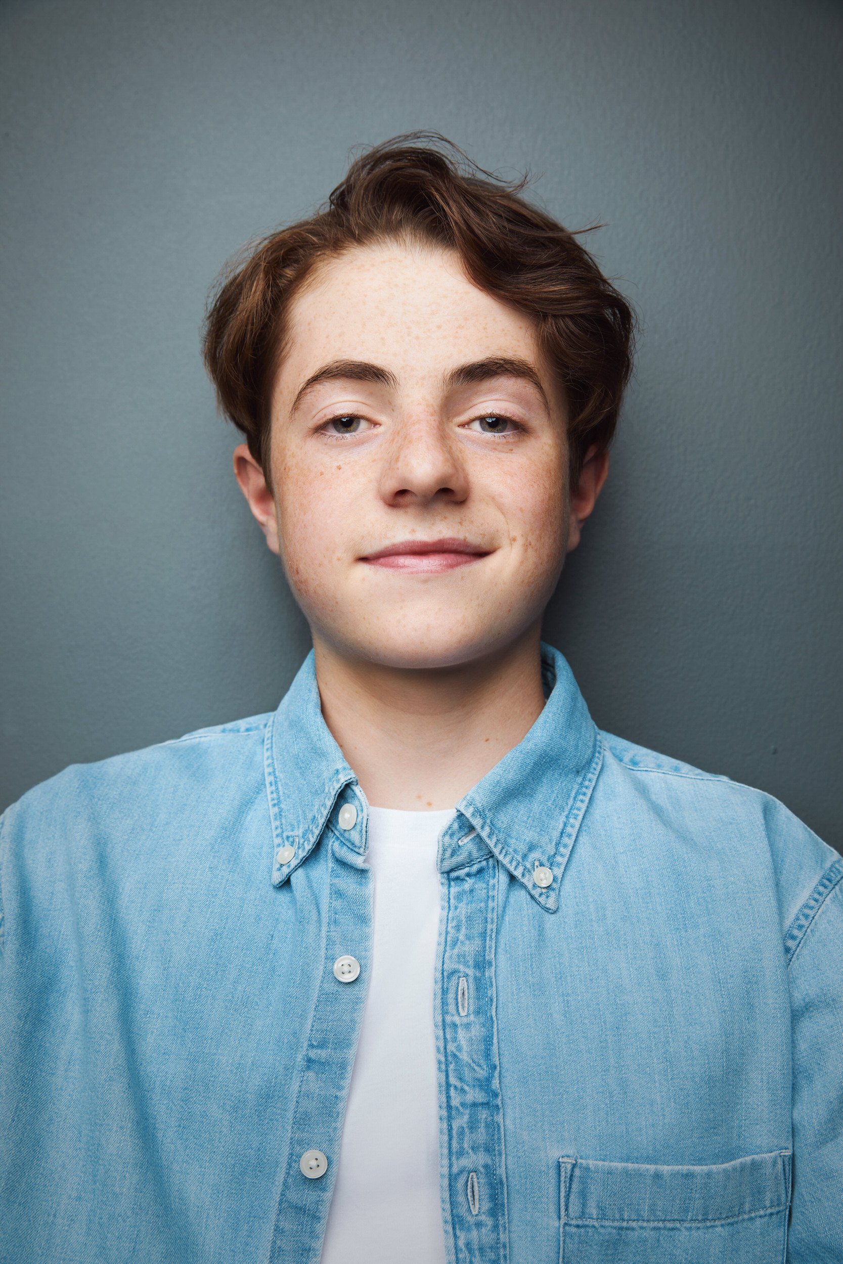 Young man with wavy brown hair and freckles in a denim shirt over a white t-shirt, smiling headshot against a grey background.