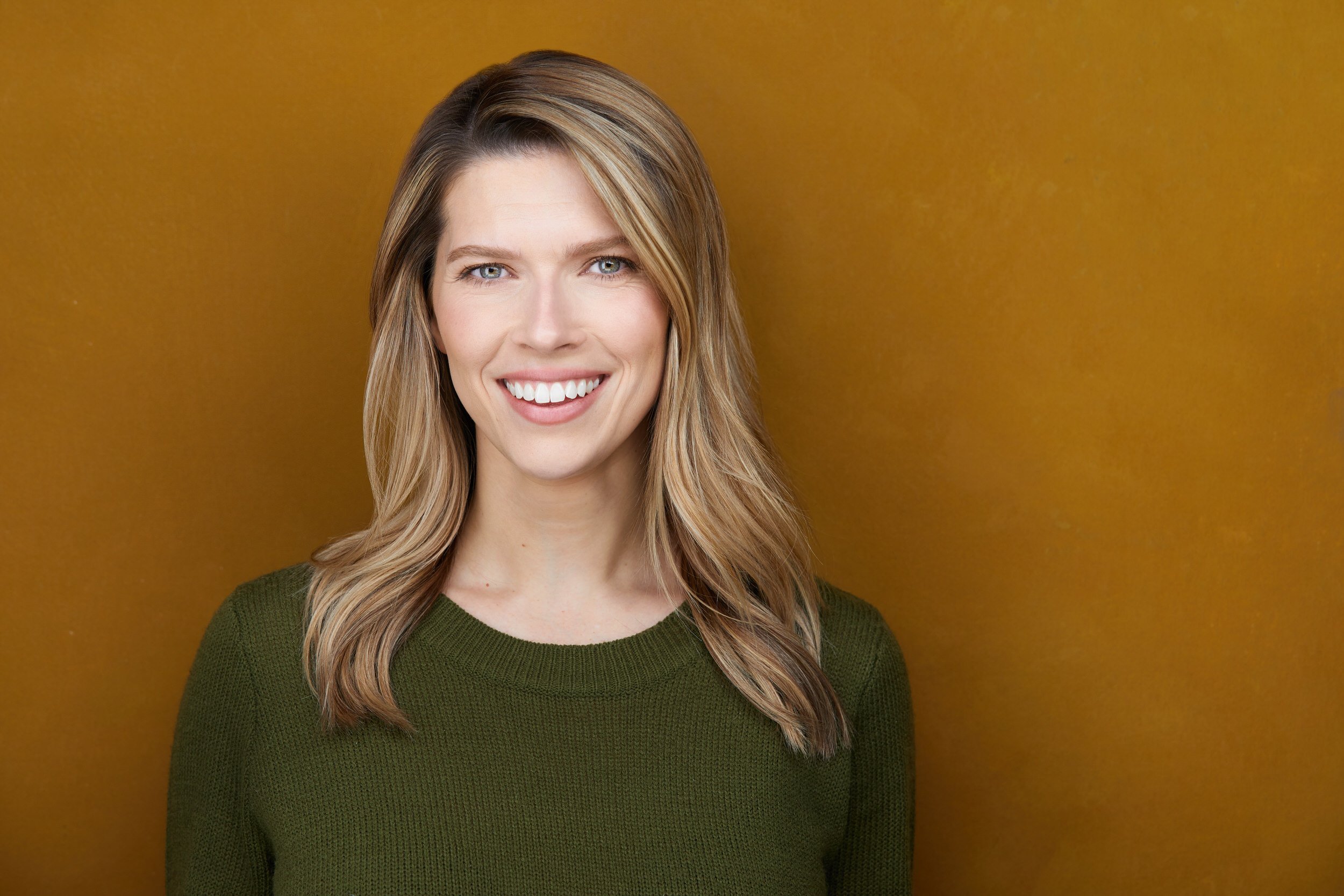 Woman with long blonde highlighted hair in an olive green sweater, smiling headshot against a mustard yellow background.