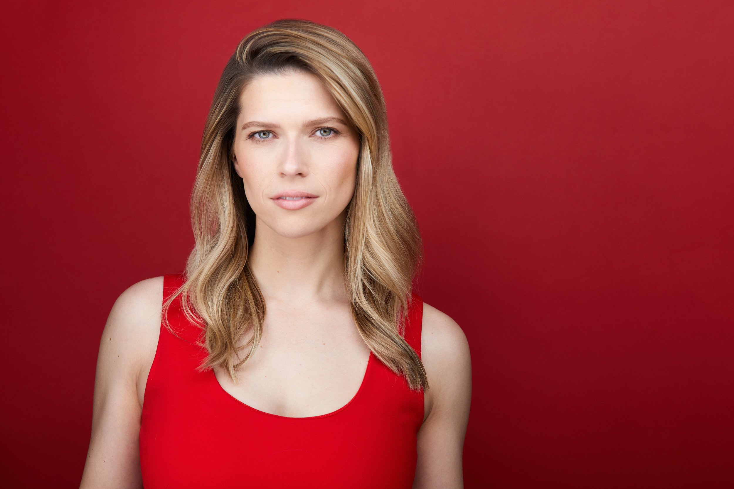 Woman with long blonde highlighted hair in a red tank top, serious headshot against a deep red background.