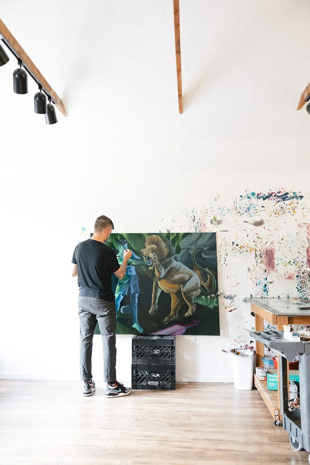 Artist painting a fantasy scene with a lion and other mythical figures in a studio with paint splatters on the wall.