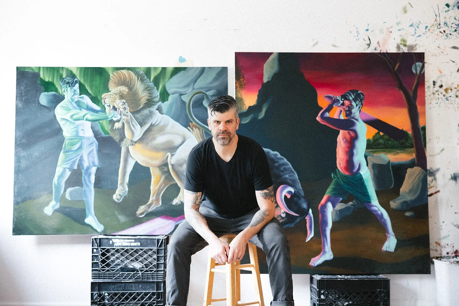 An artist with tattoos sits on a stool in front of two large colorful paintings depicting mythological scenes, in an art studio with a white wall