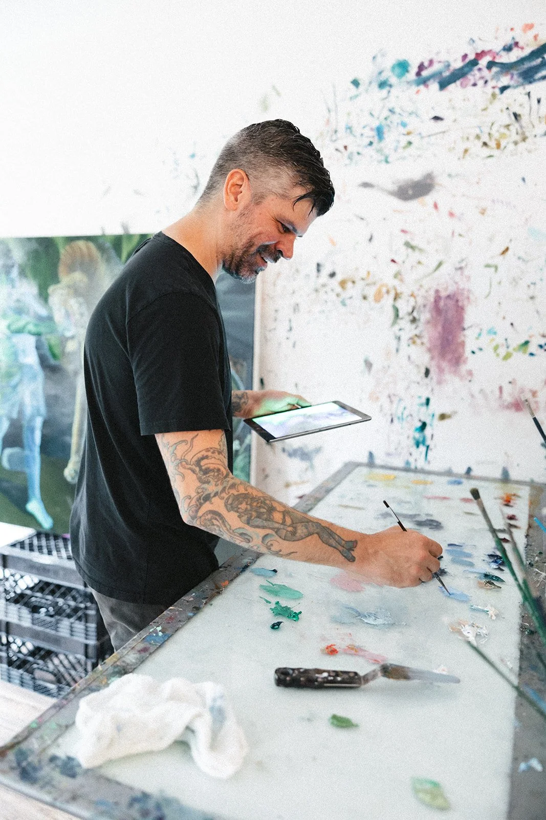 A man with tattoos on his arm, wearing a black t-shirt, is painting on a canvas with a palette knife in an art studio. He is looking at a tablet in his other hand, standing at a table with paint on it, and there is colorful splatter paint on the wall behind him.