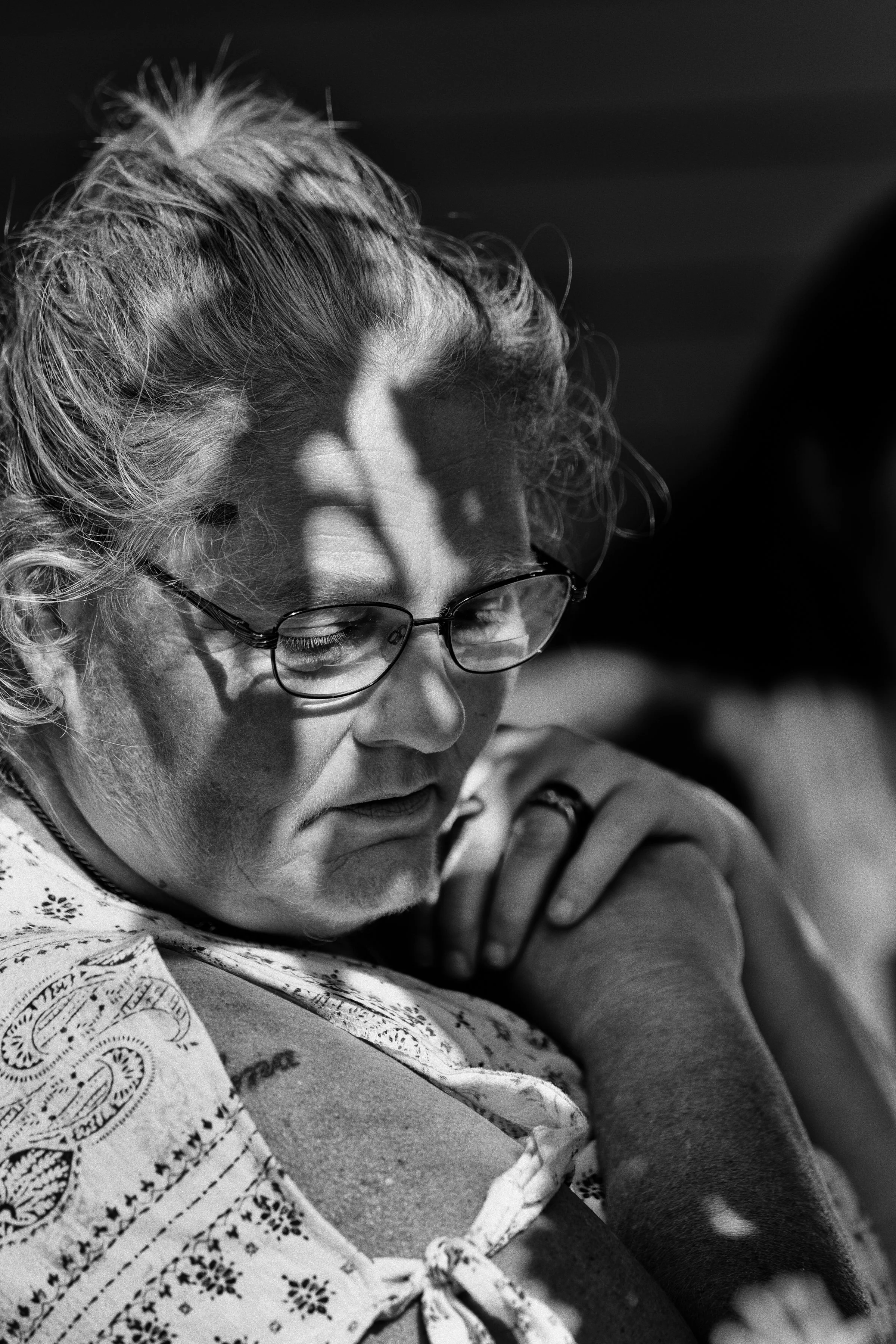 A black and white photo of a woman with glasses, resting her head on her arm, with sunlight creating shadows on her face.