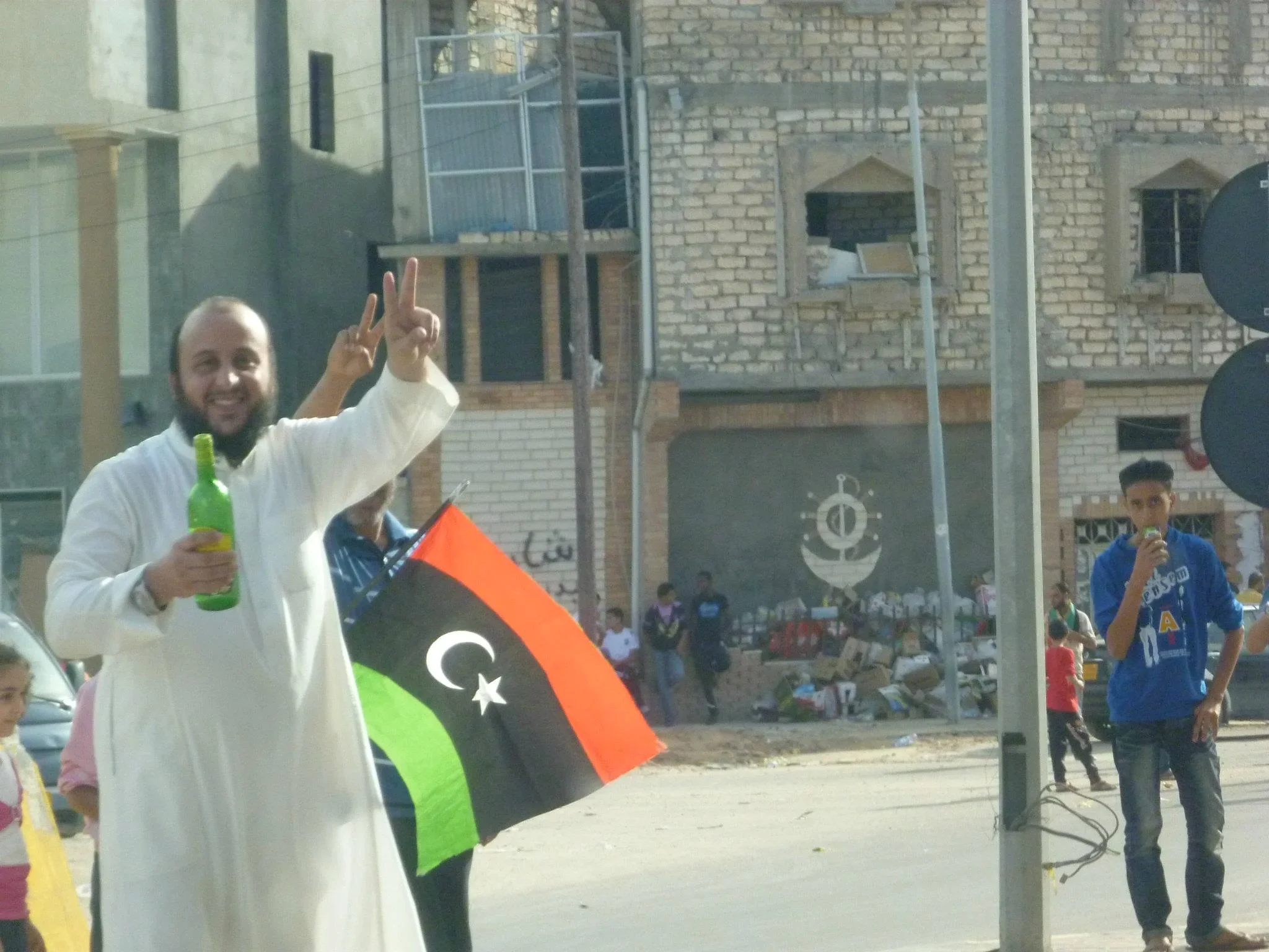 Man in a white robe with a Libyan flag and victory sign — Tripoli, 20 October 2011