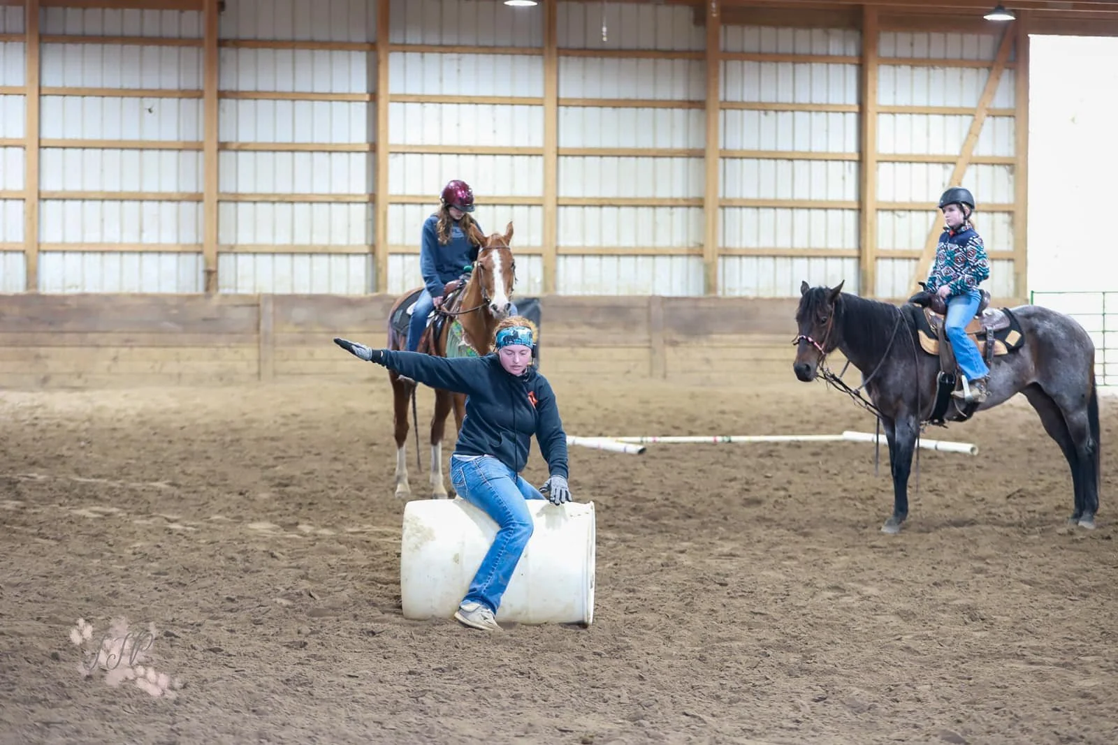 An indoor riding arena with three people and two horses. One person on a horse is riding, another person on a horse is observing, and a third person is sitting on a barrel, gesturing with an arm extended.