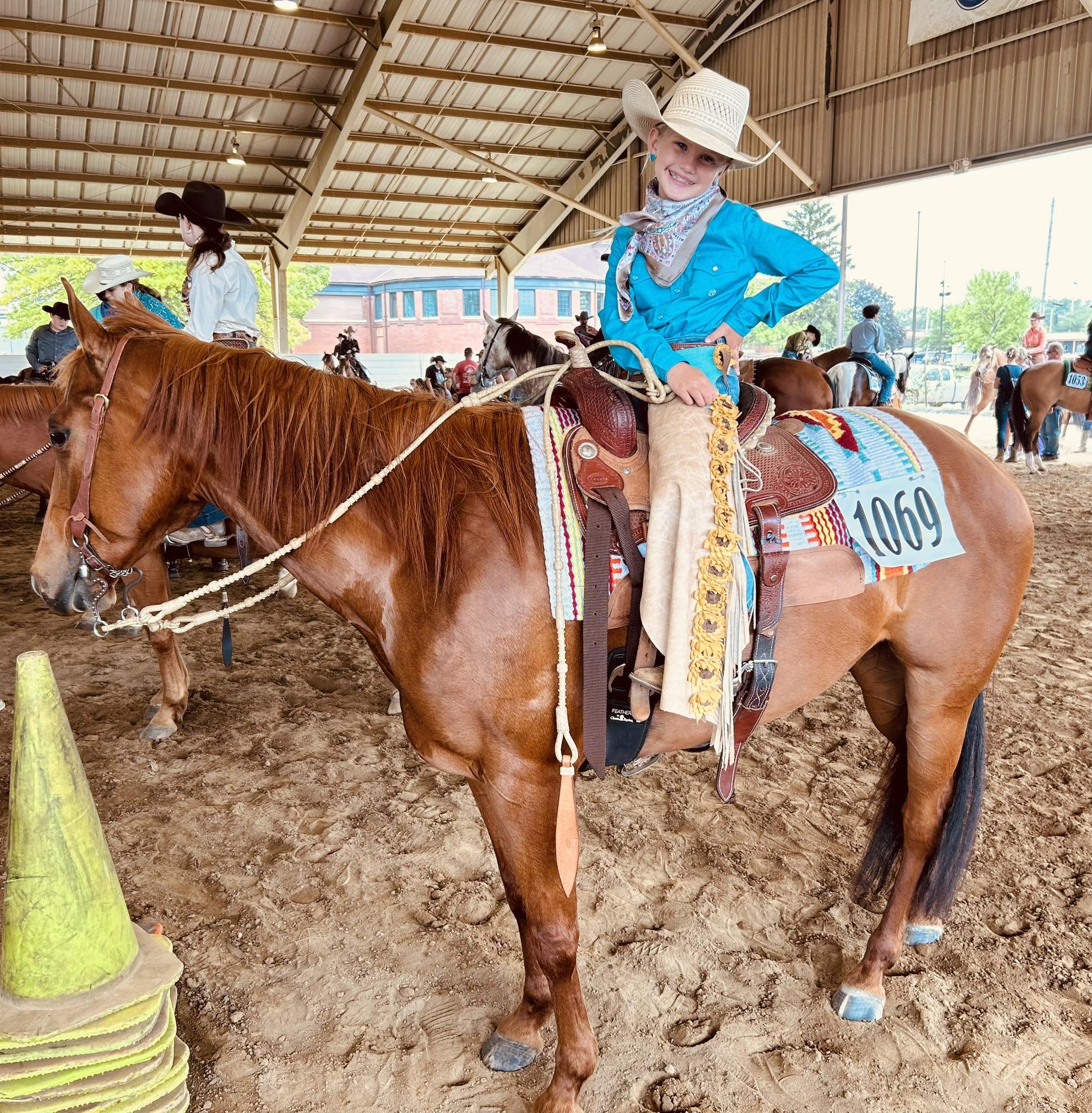 A young girl in a blue shirt and cowboy outfit is sitting on a horse, smiling, in an indoor riding arena with other riders and horses in the background.