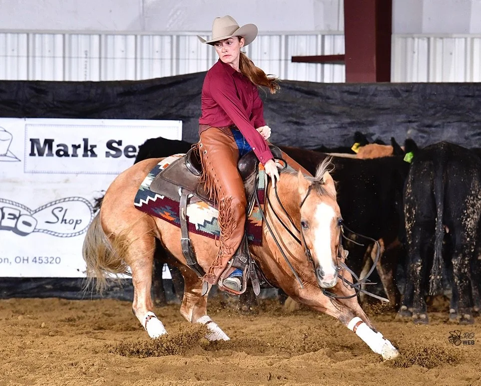 A woman wearing a cowboy hat and red long-sleeve shirt rides a galloping horse in an indoor arena. The horse has a white face blaze and white socks. There are other cattle in the background and a sign that says "Mark S...".