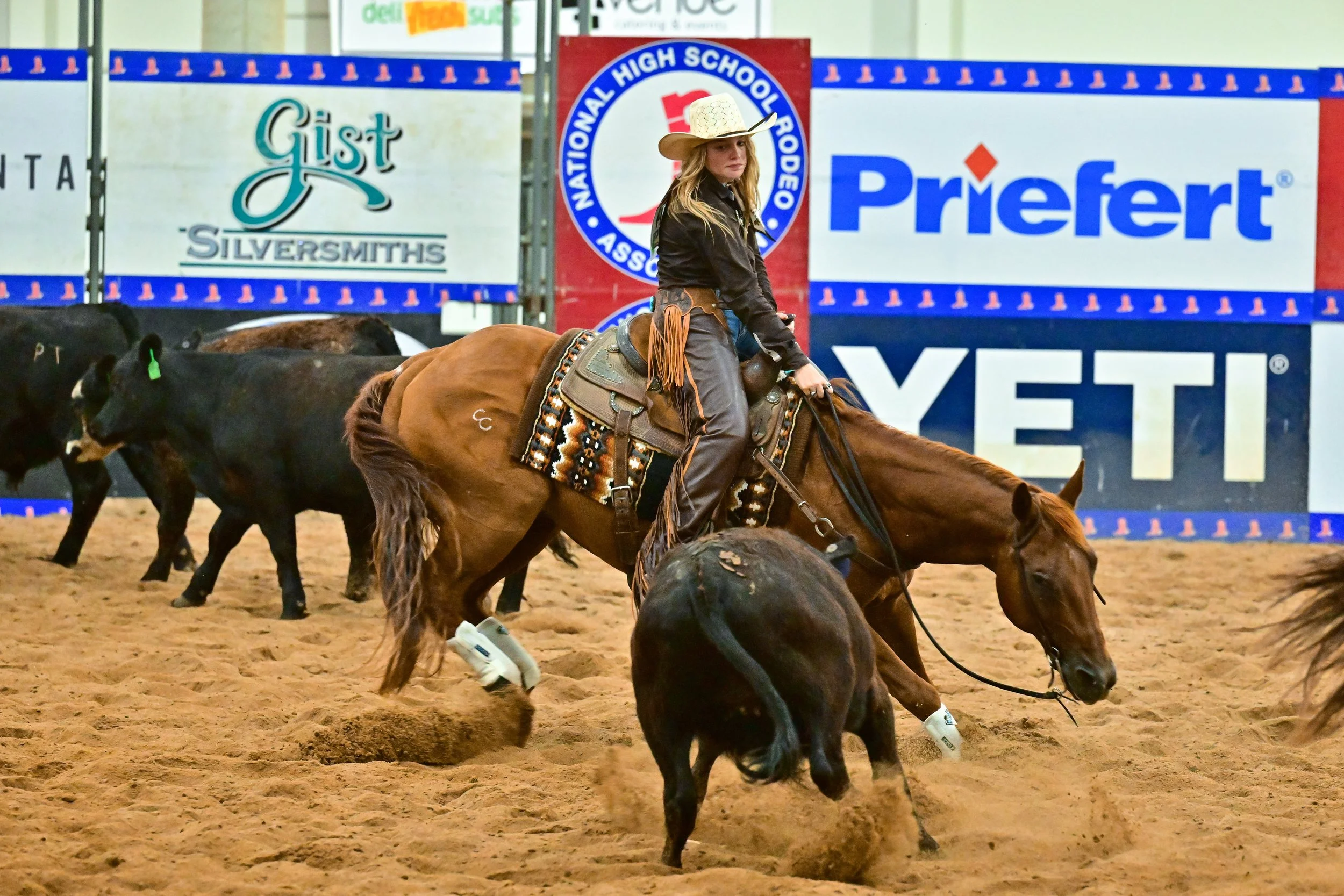 A woman dressed as a cowboy riding a brown horse at a rodeo, with cattle and a large advertising banner in the background.