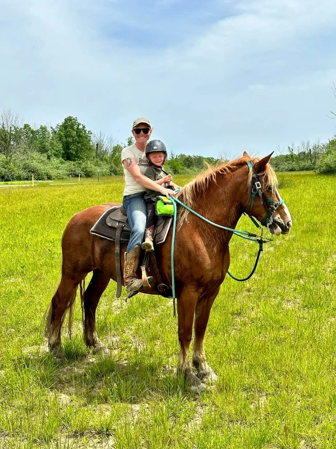 A woman and a young boy riding a brown horse in a grassy field, both wearing helmets. The woman is smiling and wearing sunglasses, while the boy is holding the reins and smiling. The field is green with trees in the background under a partly cloudy s
