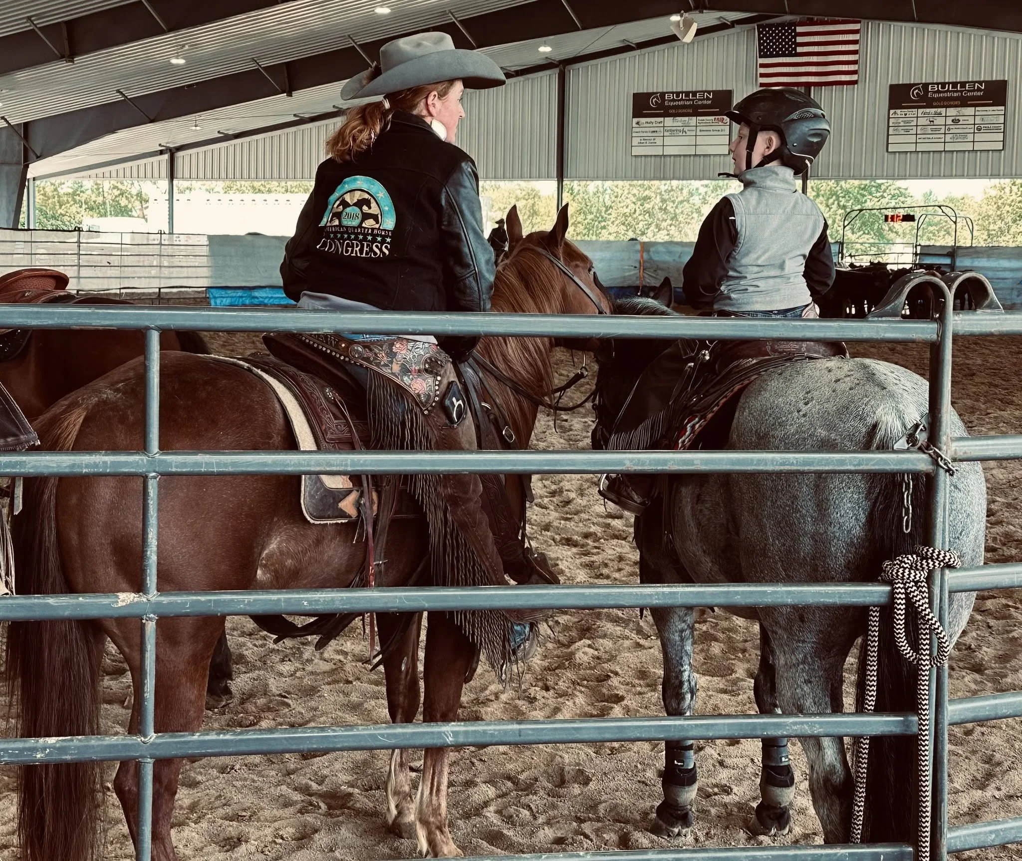 Two people on horseback inside a covered riding arena, with a large American flag hanging on the wall behind them.