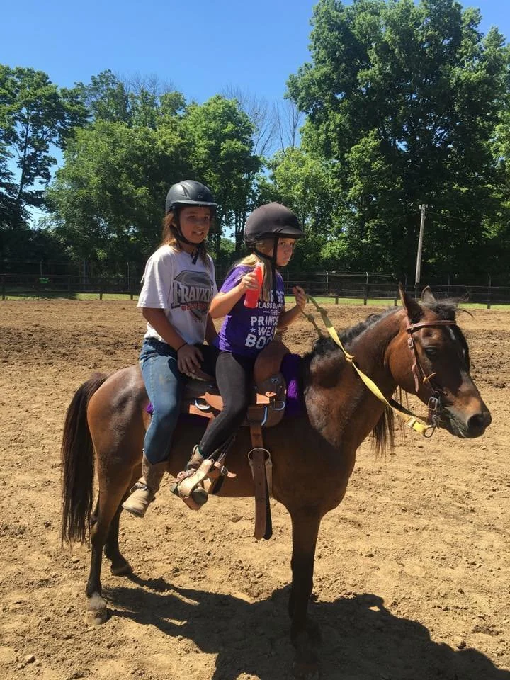 Two young girls wearing helmets riding a brown horse in an outdoor riding arena with trees and a clear blue sky in the background.