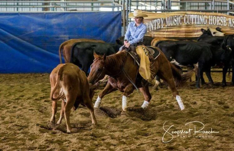 A woman in cowboy hat riding a horse in an indoor arena with calves and cows and a banner that reads "Country Music Cutting Horse Association."