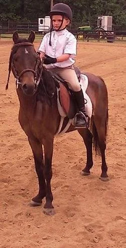 A young girl wearing a riding helmet, white shirt, and riding pants sits on a horse in an outdoor riding arena.