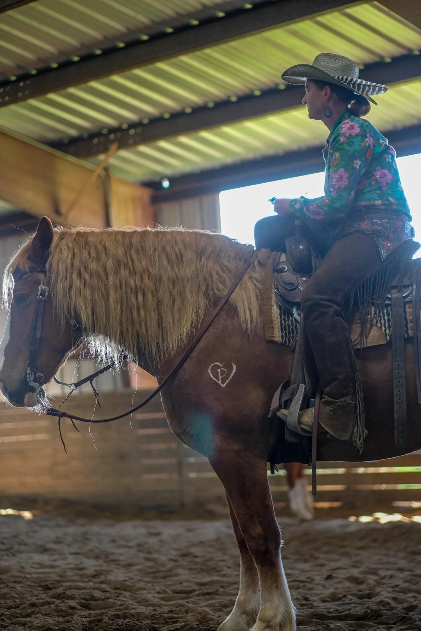 A woman wearing a large straw hat, colorful floral shirt, and turquoise earrings is riding a light brown horse inside a barn with a metal roof. The horse's head and part of its body are visible, and the woman is holding the reins, sitting on a saddle