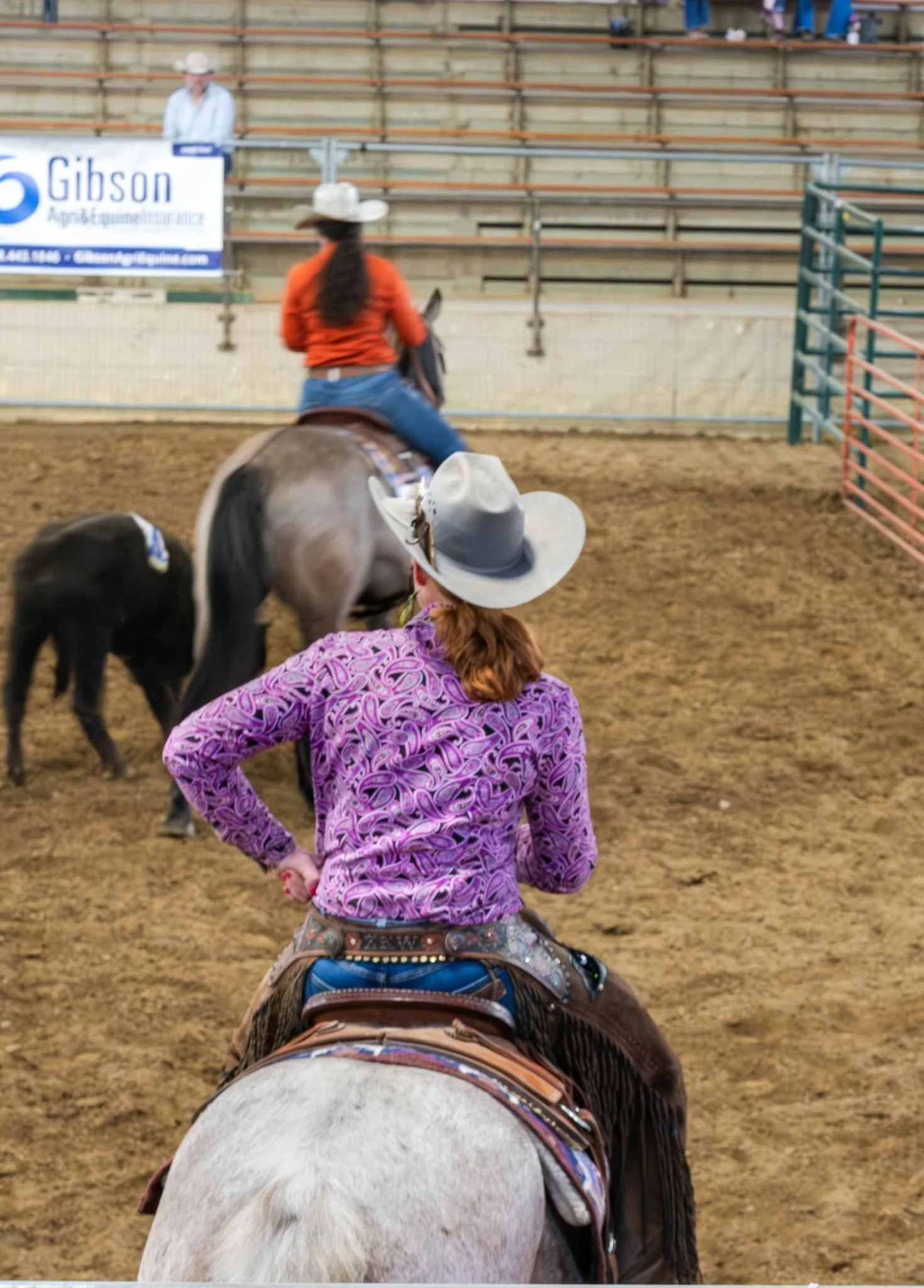 A person riding a horse in an indoor arena, wearing a white cowboy hat, purple shirt, and blue jeans, with hands on hips; another person on a horse is in front, wearing a patterned shirt and cowboy hat.