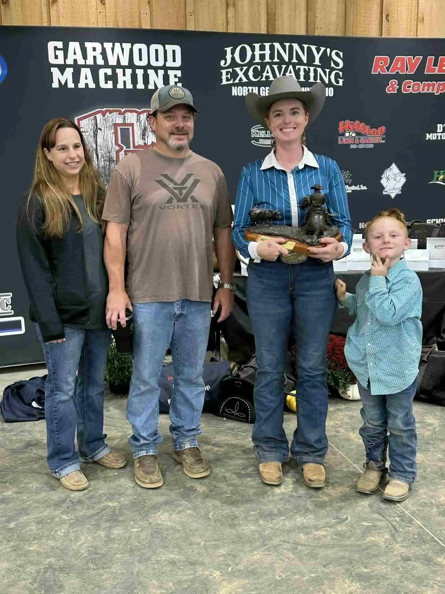 Group of people standing indoors in front of a black backdrop with various logos and text, including "Garwood Machine" and "Johnny's Excavating." One woman is holding a small statue. The group includes two women, two men, and a young boy, all dressed casually.