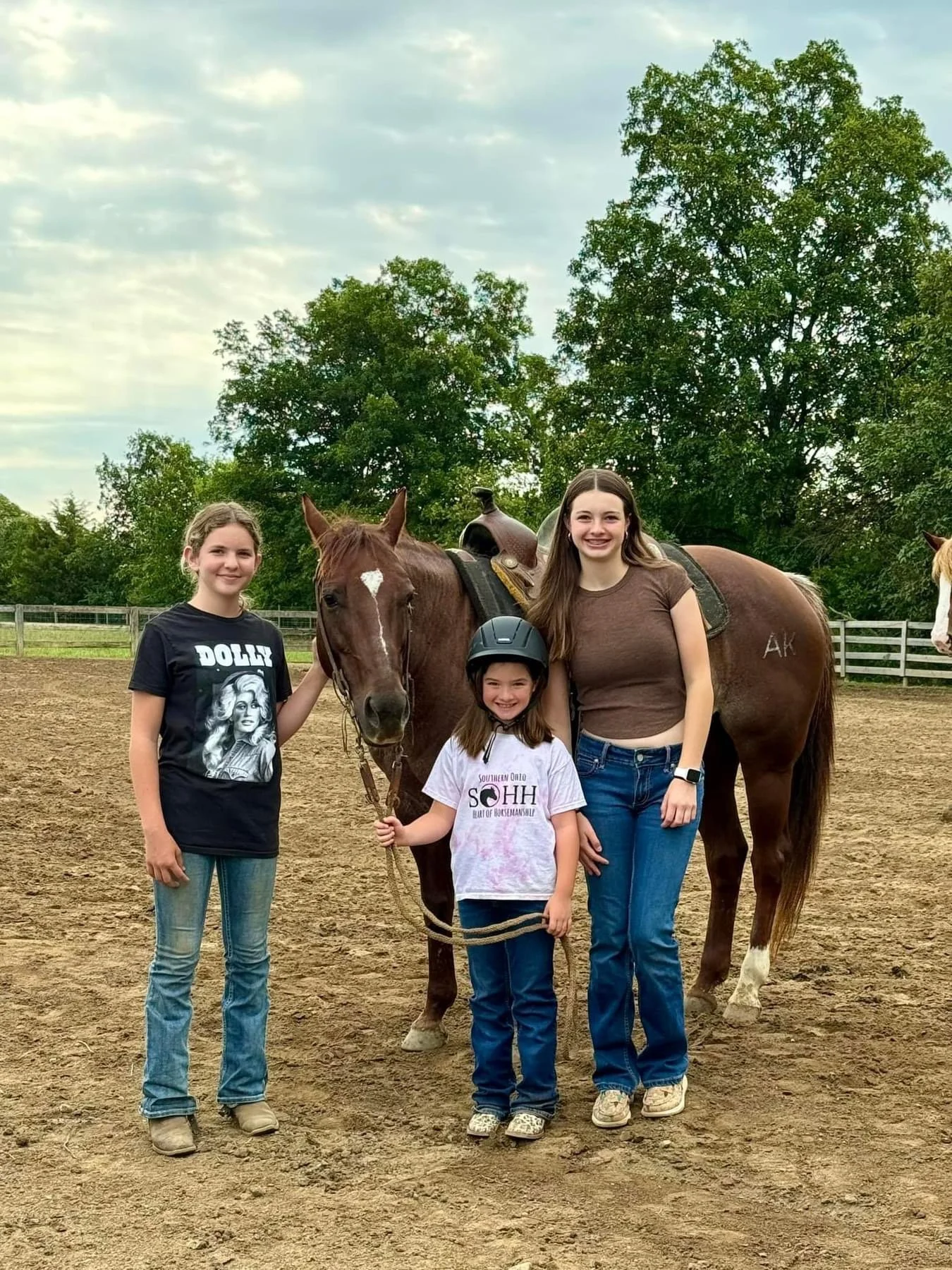 Three girls and a brown horse in a fenced outdoor riding area with trees in the background. The girls are smiling and standing close to the horse, which has a saddle and a bridle.