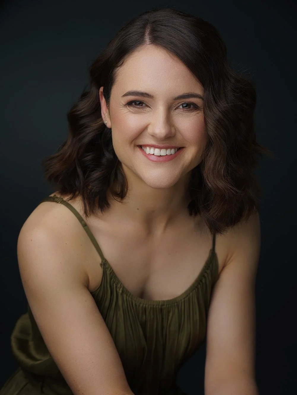 A woman with shoulder-length dark wavy hair, smiling, wearing a sleeveless green top, against a dark background.
