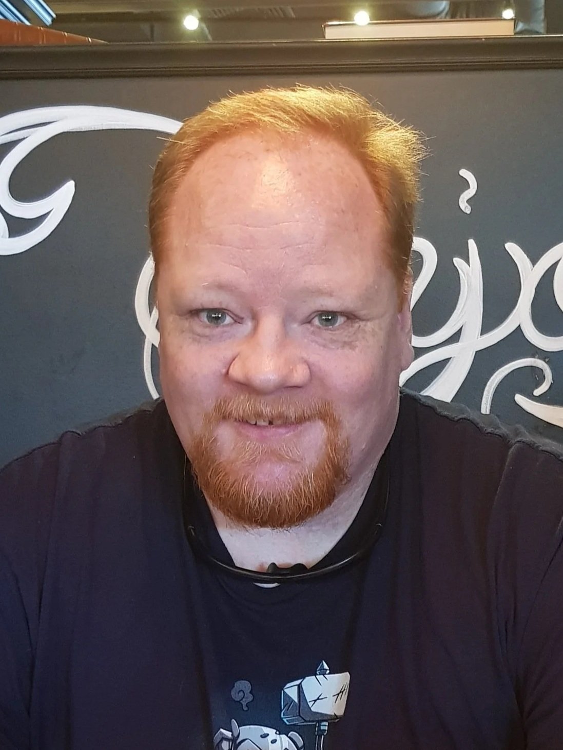 Close-up of a man with red hair, freckles, and a beard, wearing a black shirt, standing in front of a background featuring tall beer glasses and bottles.