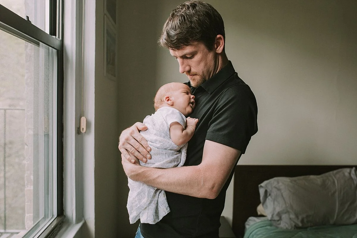 nyc family and newborn photographer dad holds sleeping newborn son in bedroom of Park Slope apartment