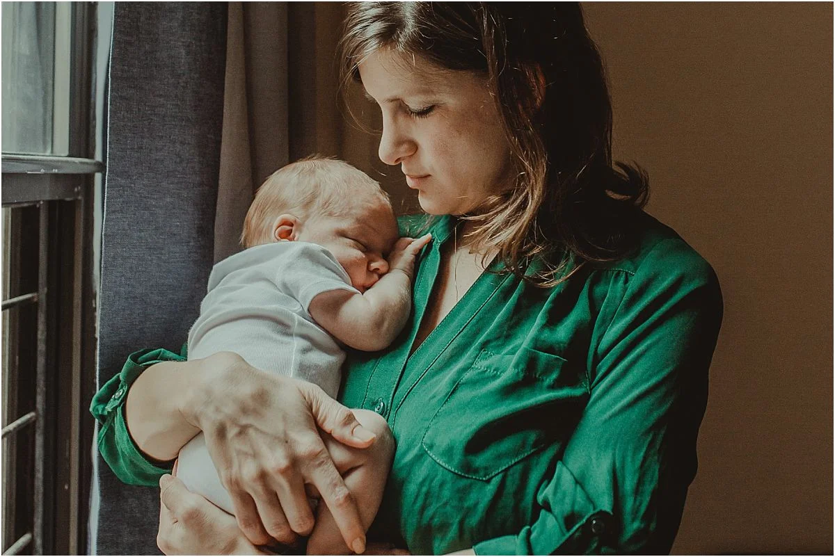 nyc family and newborn photographer mom holds sleepy newborn son near light filled window