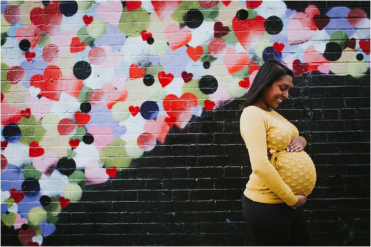 nyc family and newborn photographer expecting mom holds belly bump in front of heart graffiti wall for maternity session