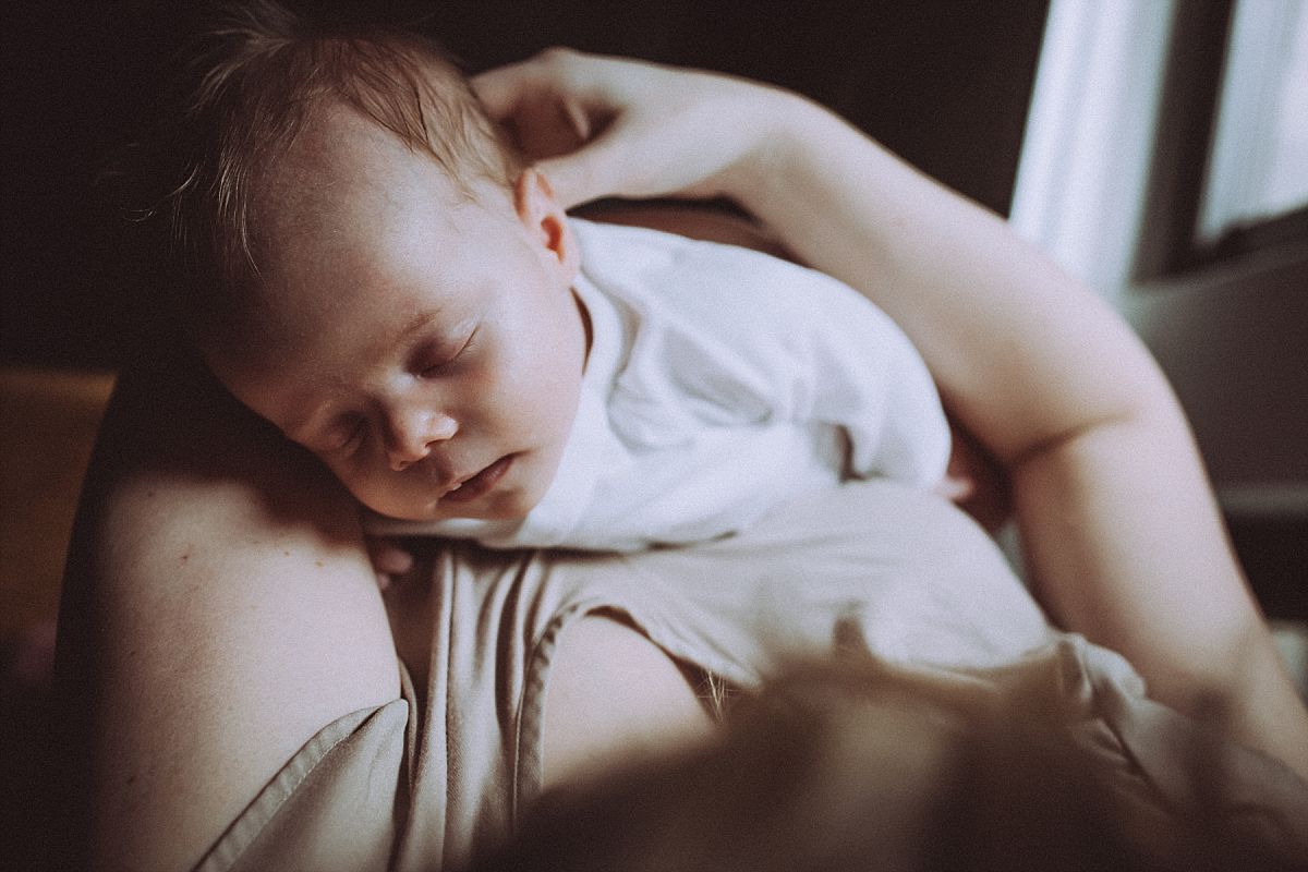 nyc family and newborn photographer new mom holds sleeping newborn baby girl in arms in front of light filled windown of Chelsea home