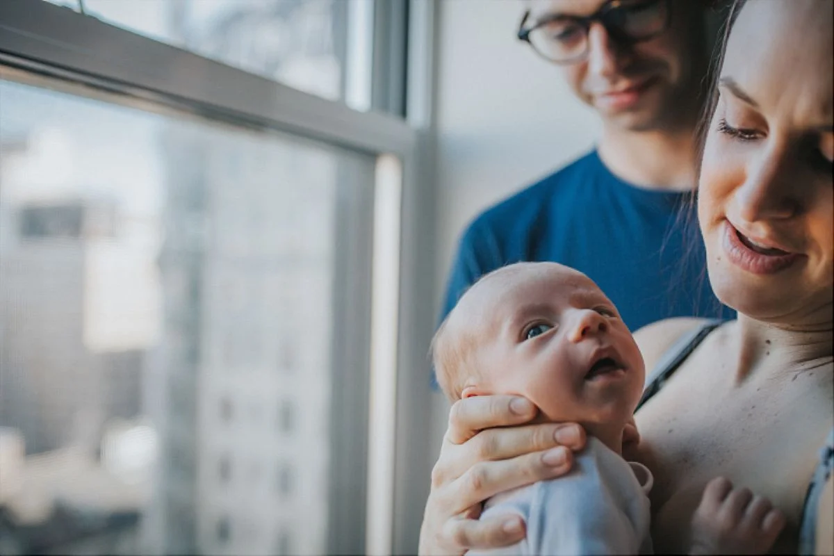 nyc family and newborn photographer mom holds newborn son by window with dad in background