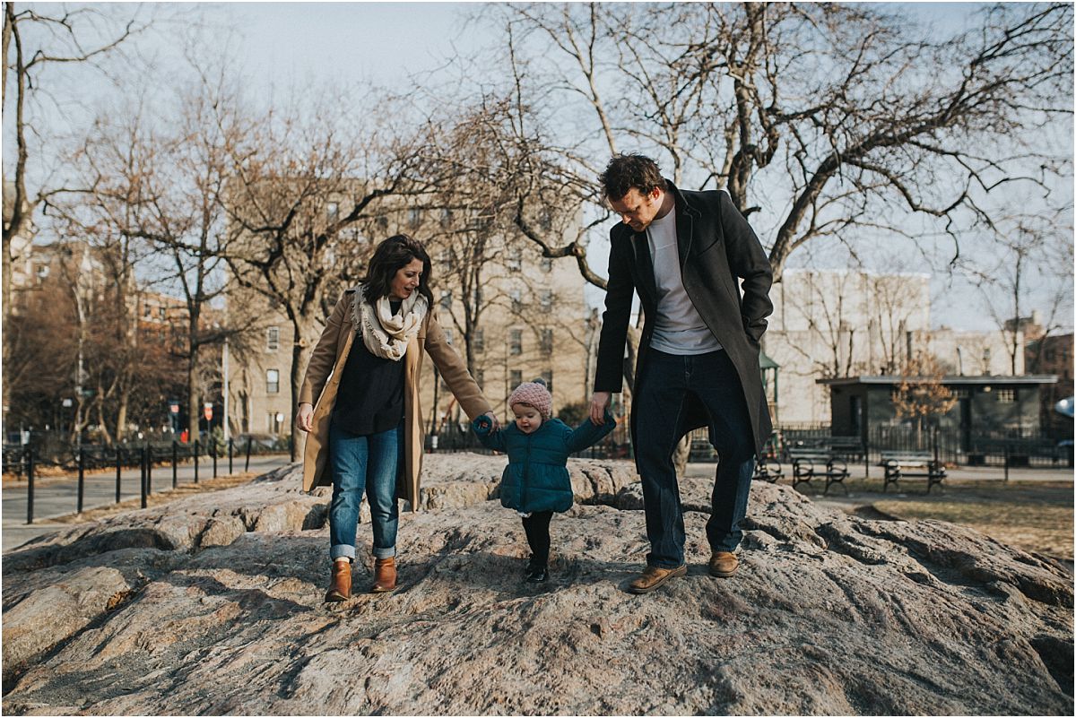 nyc family and newborn photographer mom, dad and daughter climbing on rock in local nyc park bench