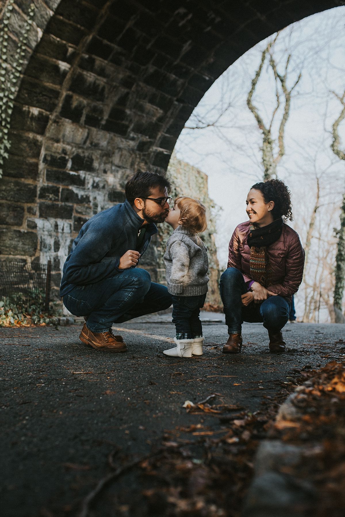 nyc family and newborn photographer mom and dad sit with toddler under bridge and toddler kisses dad in fun outdoor family session