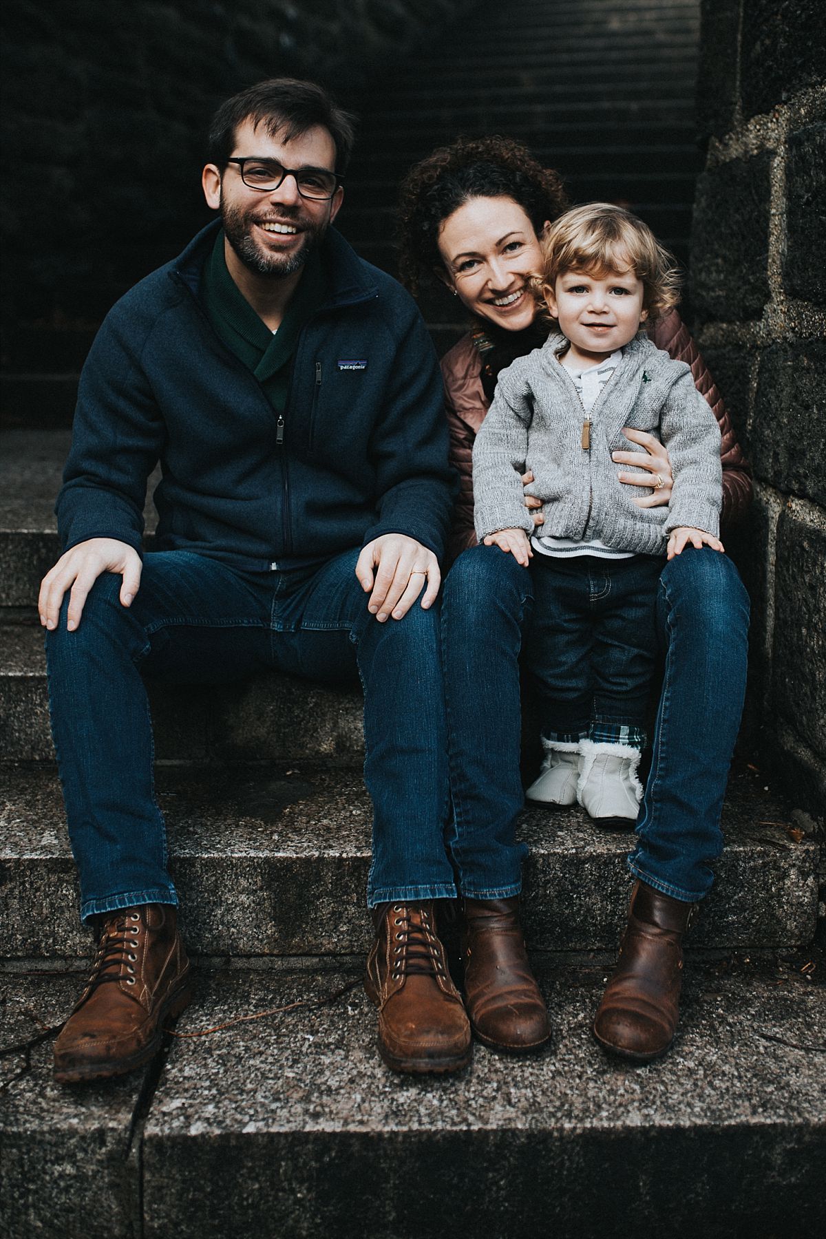 nyc family and newborn photographer mom and dad sit with toddler under bridge and toddler kisses dad in fun outdoor family session