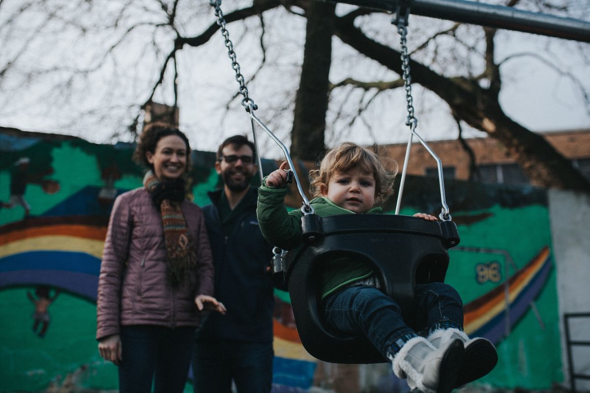 nyc family and newborn photographer mom and dad push toddler on swing at nyc playground in outdoor winter family session