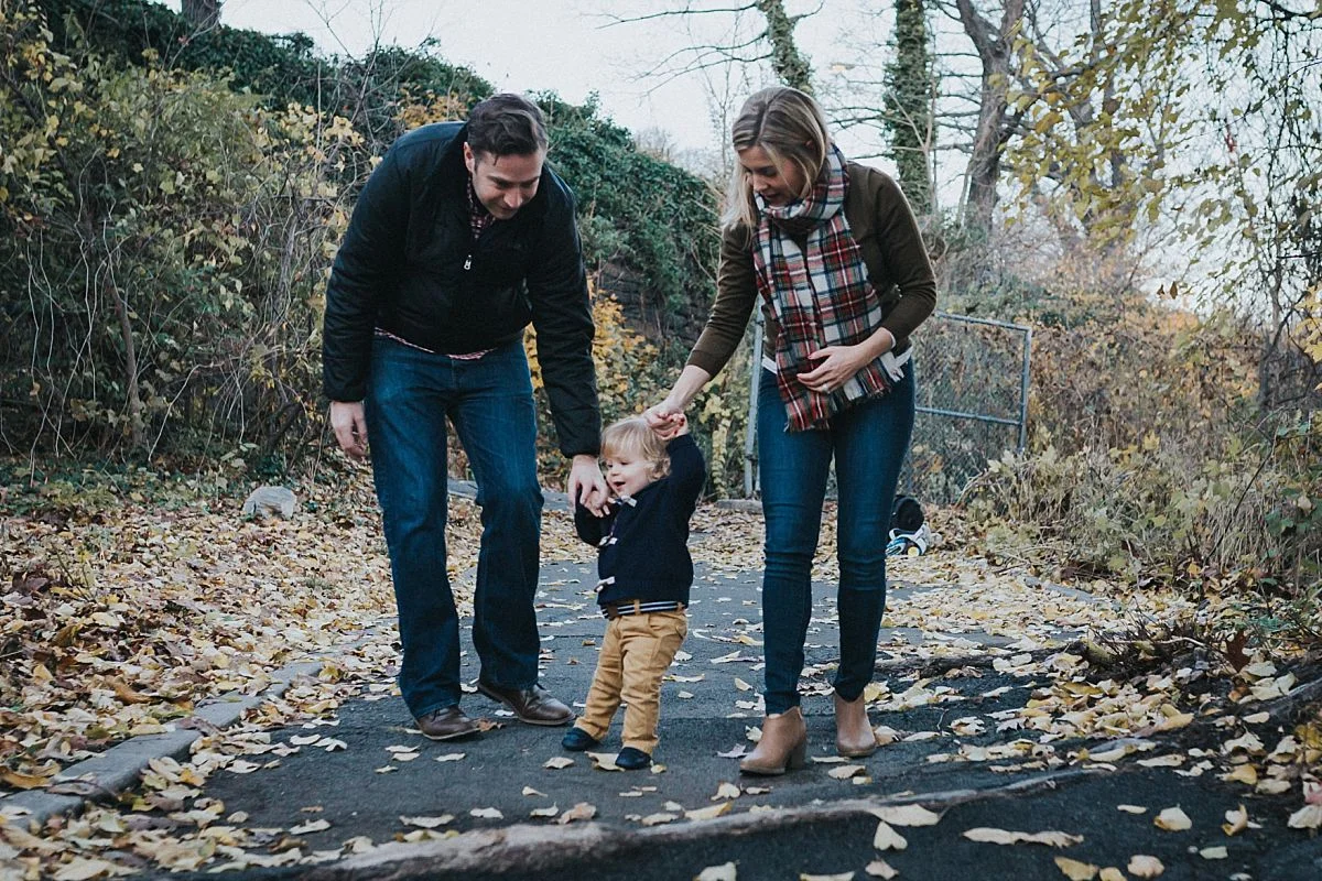 nyc family and newborn photographer mom, dad and toddler boy walk amongst fall leaves in nyc park for family lifestyle session