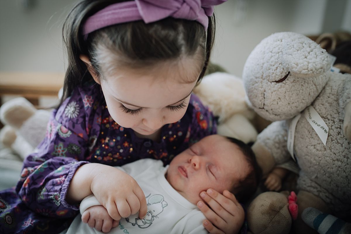 nyc family and newborn photographer big sister holds newborn sister while sitting on bed with stuffed animals