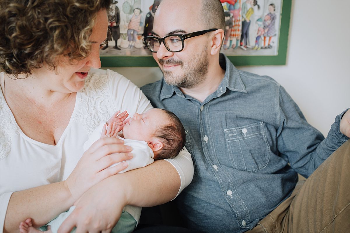 nyc family and newborn photographer mom and dad sit on bed holding newborn girl while newborn sleeps peacefully