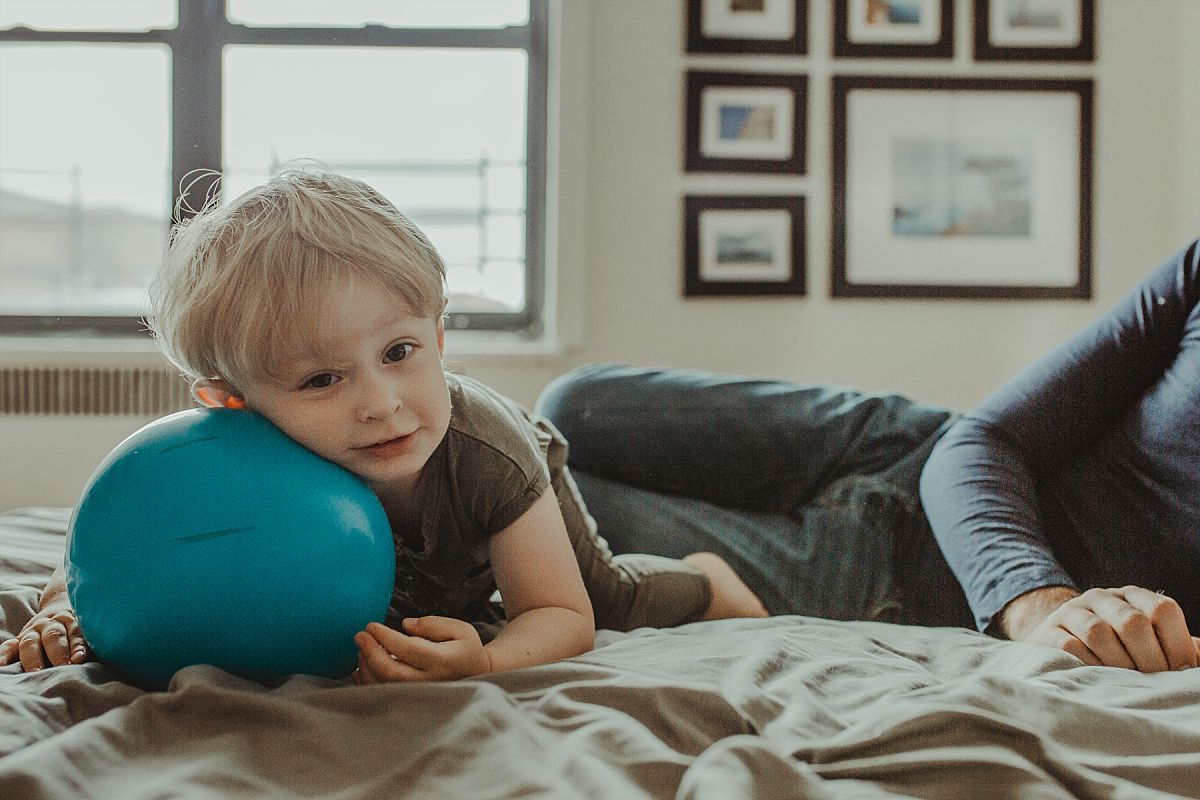 nyc family and newborn photographer boy plays with big blue ball on parents bed in nyc apartment