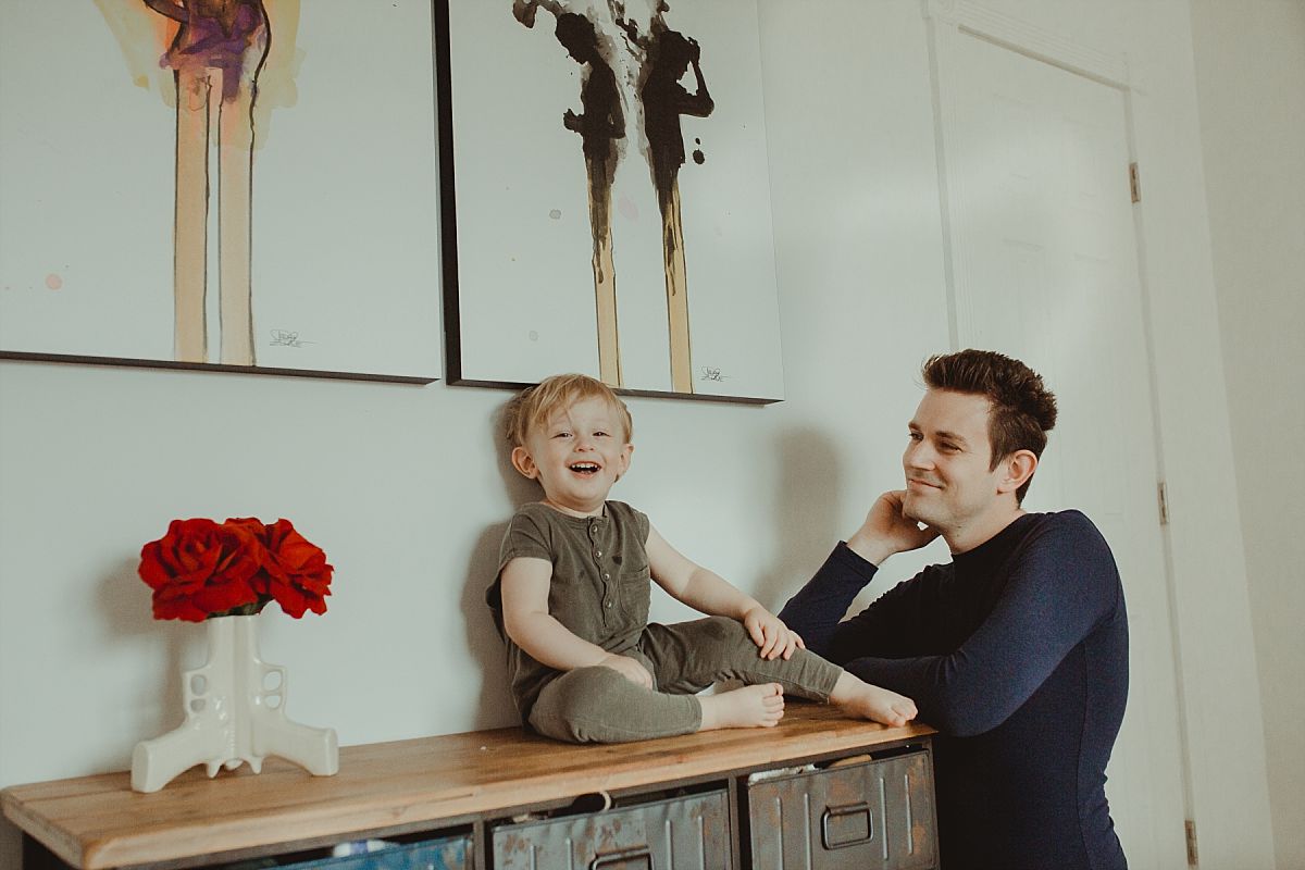 nyc family and newborn photographer sons sits on chester draws giggling while dad looks on lovingly