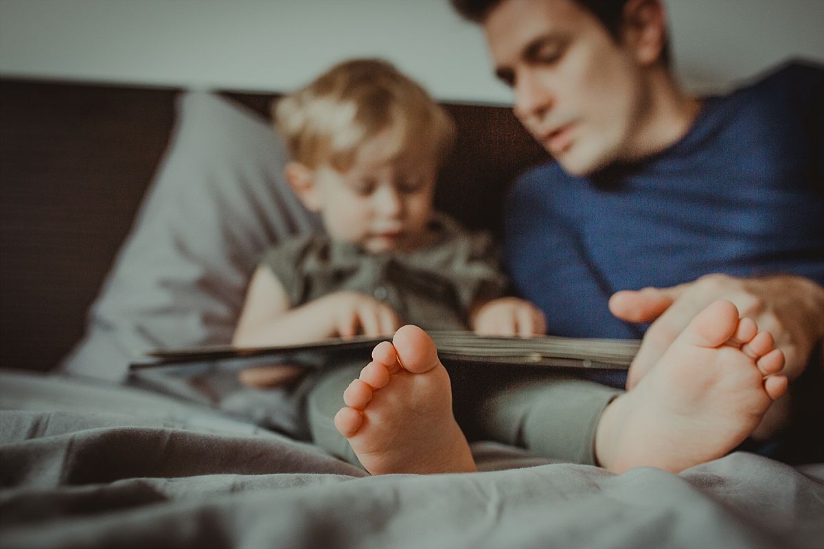 nyc family and newborn photographer mom and dad enjoy reading book on bed at nyc in home session with close up of toddler feet
