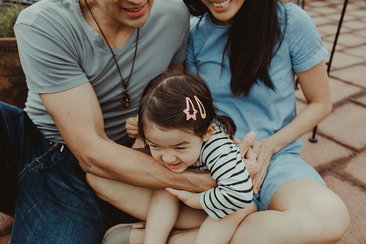 daughter enjoys tickles from mom and dad while sitting on mom's lap on rooftop during balmy afternoon photo session