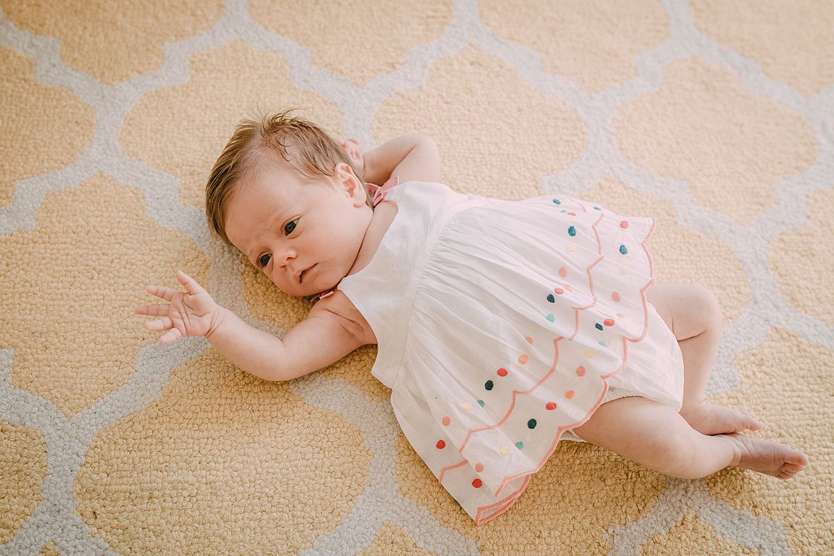 newborn baby girl with outstretched arm lying on yellow and white rug in cute polka dot newborn dress