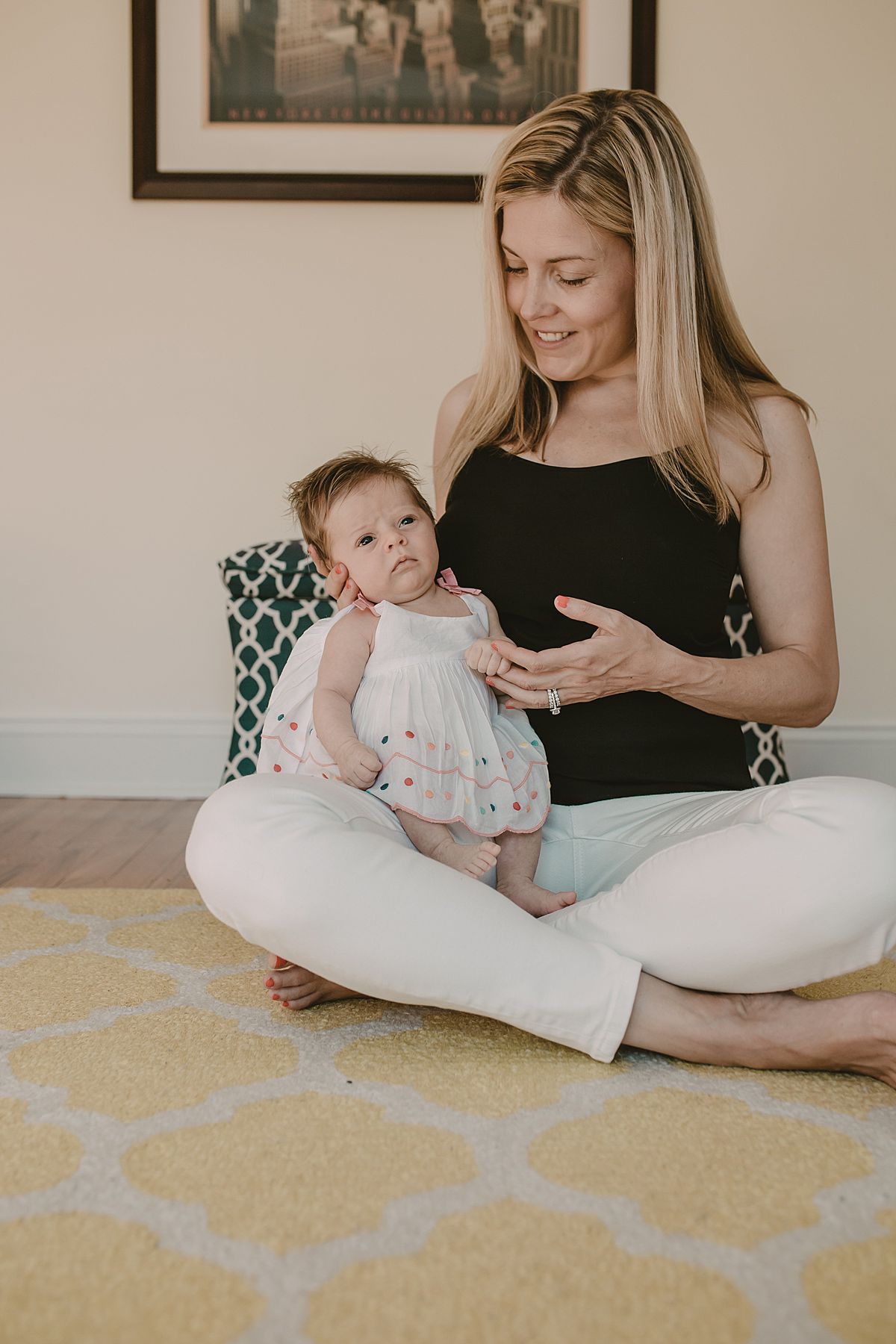 mom and newborn girl sit on yellow and white rug in kids room. images taken during family nyc family lifestyle session by krystil mcdowall photography