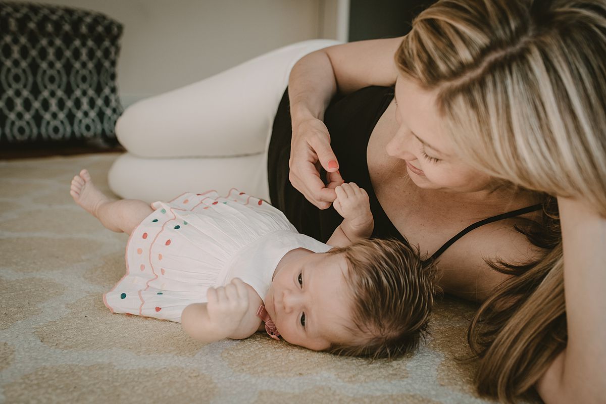 mom and daughter laying on while and yellow rug while mom plays with daughter's hand and smiles lovingly at daughter