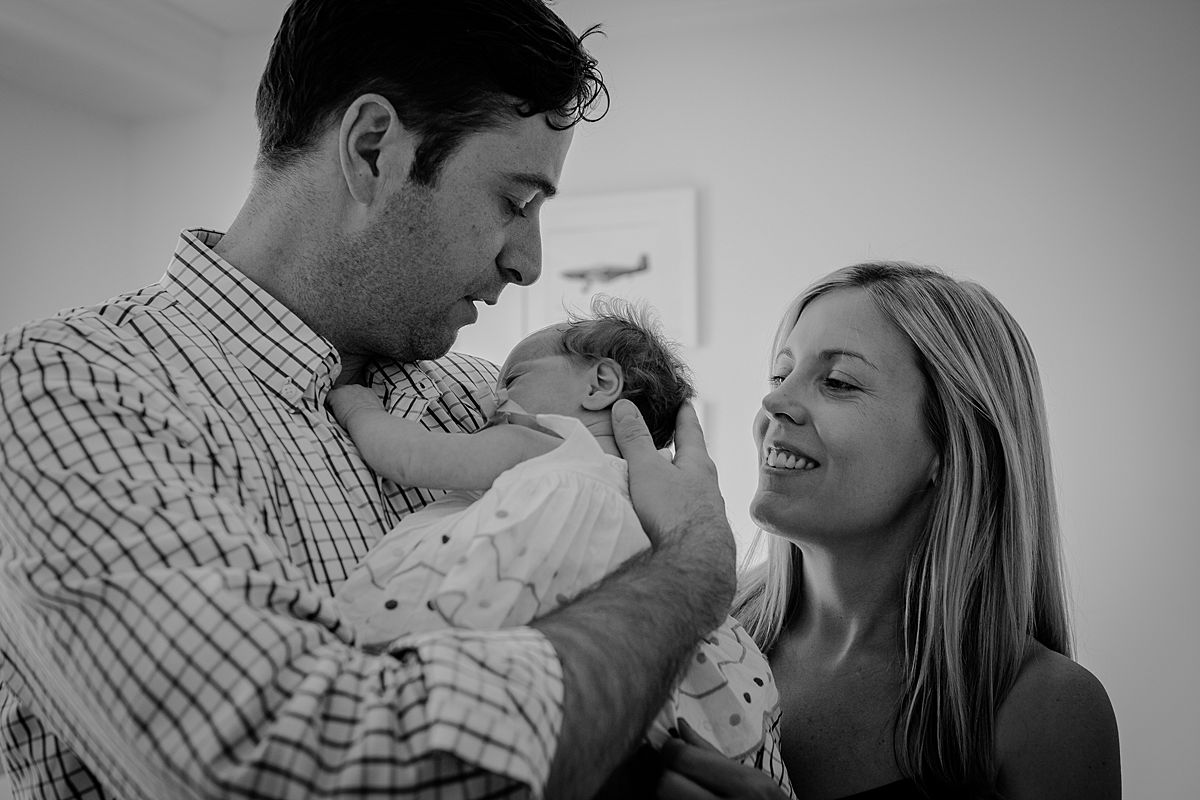 mom and dad looking at newborn daughter with lots of love and smiles andlots of backlight streaming through window. nyc newborn session by krystil mcdowall