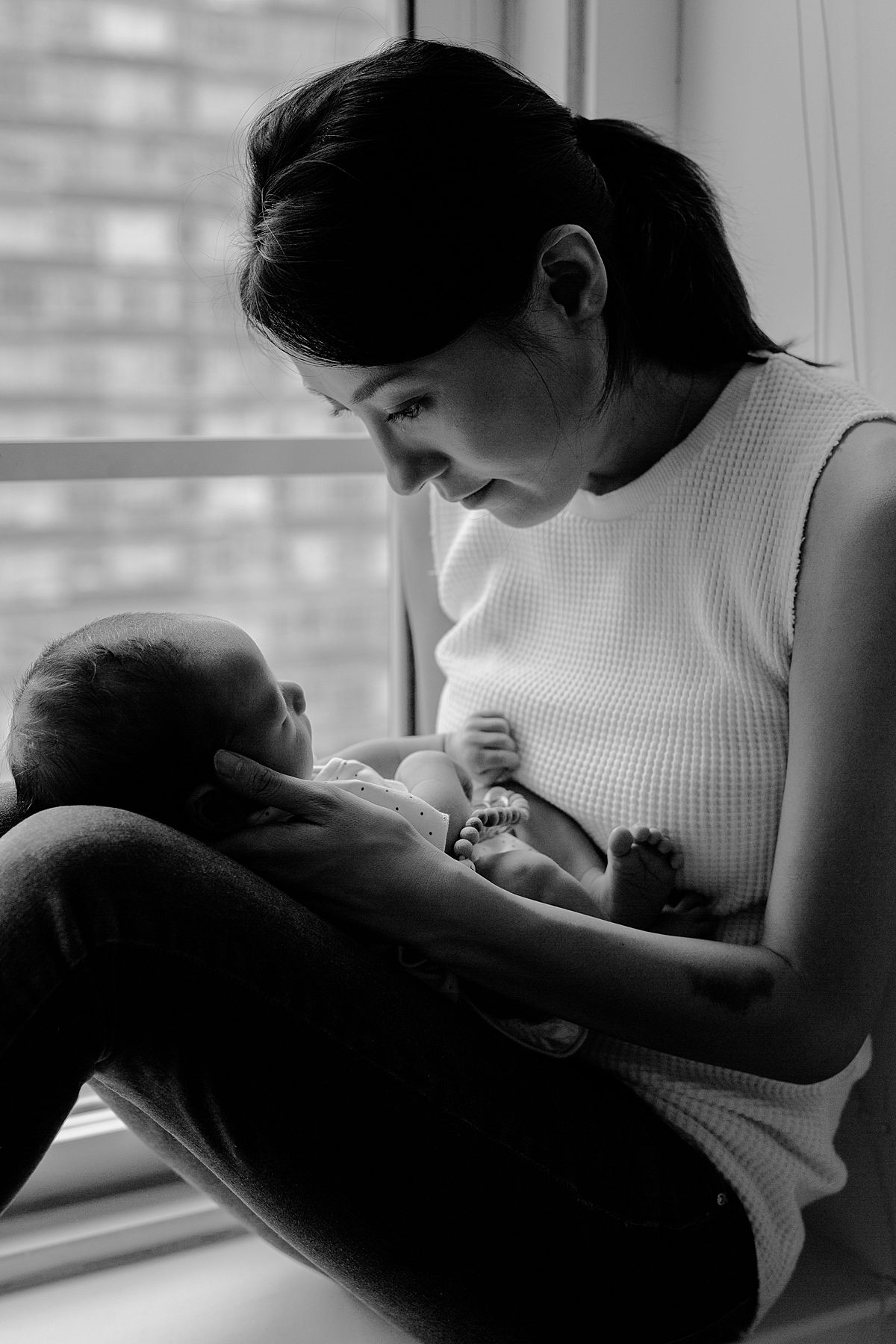 black and white image of mom holding newborn son in lap while sitting on dun drenched window sill. image taken in nyc apartment by nyc family and newborn photographer krystil mcdowall