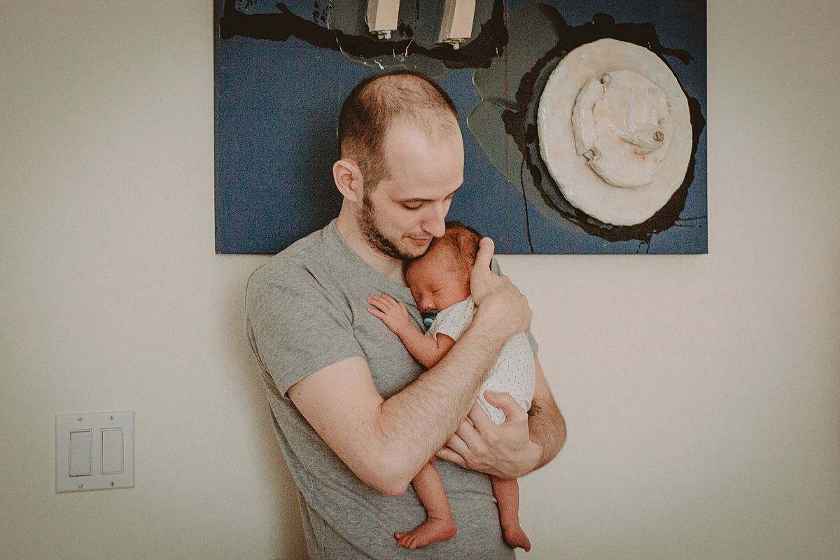 dad holds newborn baby son across chest while he sleeps. dad stands in front of funky wall hanging