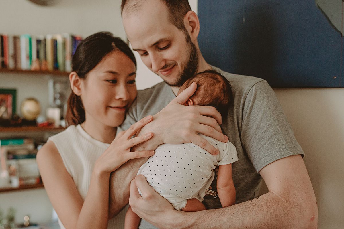 dad holds newborn baby son while mom lays hand on son't back and. family stands in front of funky wall hanging in nyc downtown apartment. image by krystil mcdowall photography