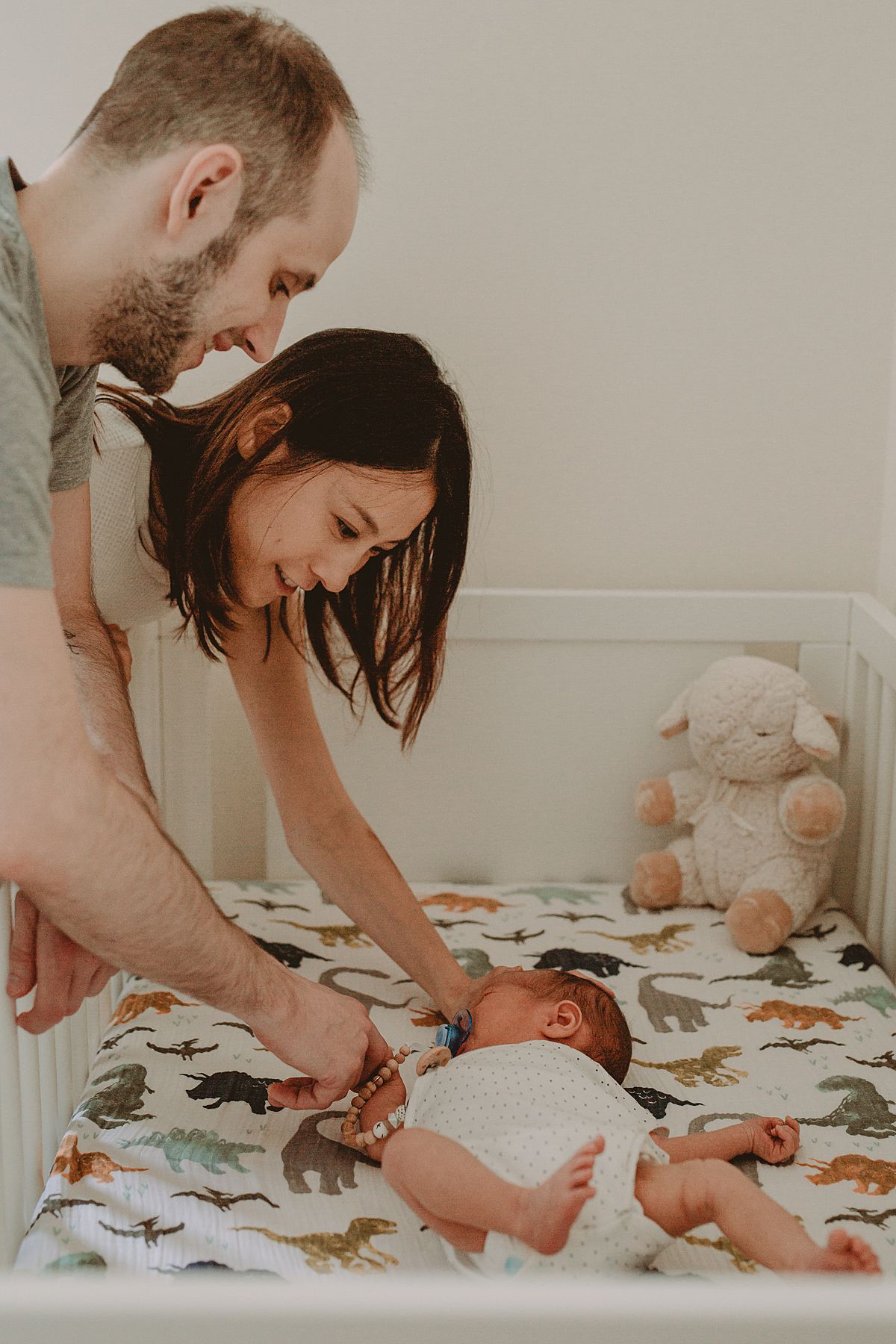 mom and dad caressing newborn as he tries to sleep in crib on his dinosaur sheets. photo by nyc family photographer krystil mcdowall