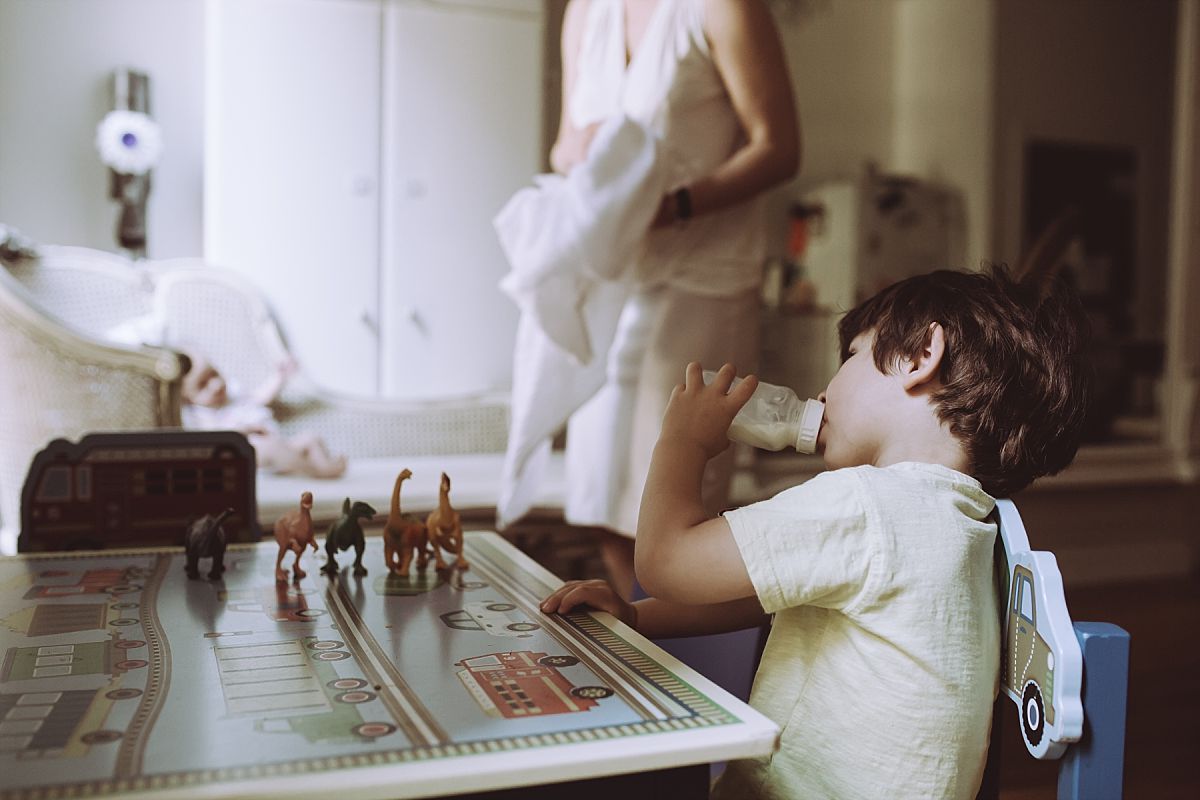 son drinking milk sitting at table with his dinosaurs overlooking mum and baby girl on chair